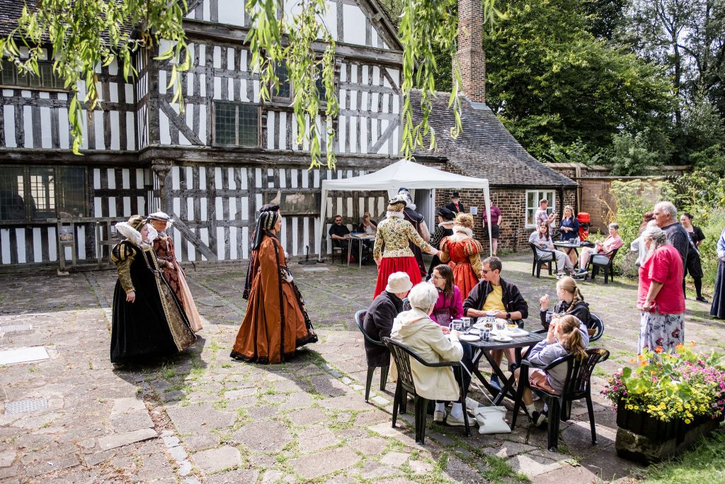 A group of people in Tudor dress walk in front of Ford Green Hall. There are people sat at tables outside having cake and drinking coffee.