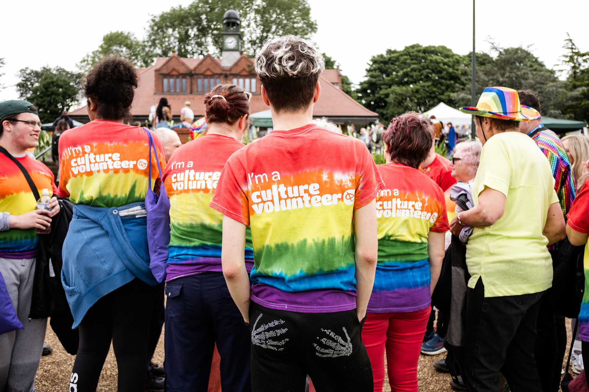 A group of volunteers stand with their backs to the photographer in Pride themed Culture Volunteer T-Shirts