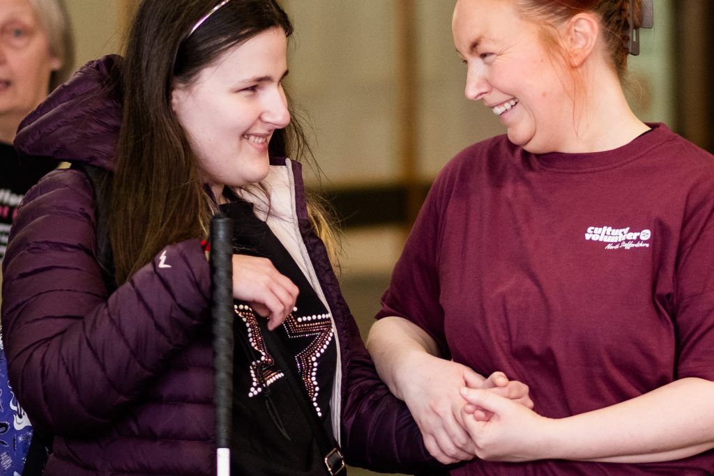 A woman in culture volunteer is smiling at another woman who is carrying a cane.